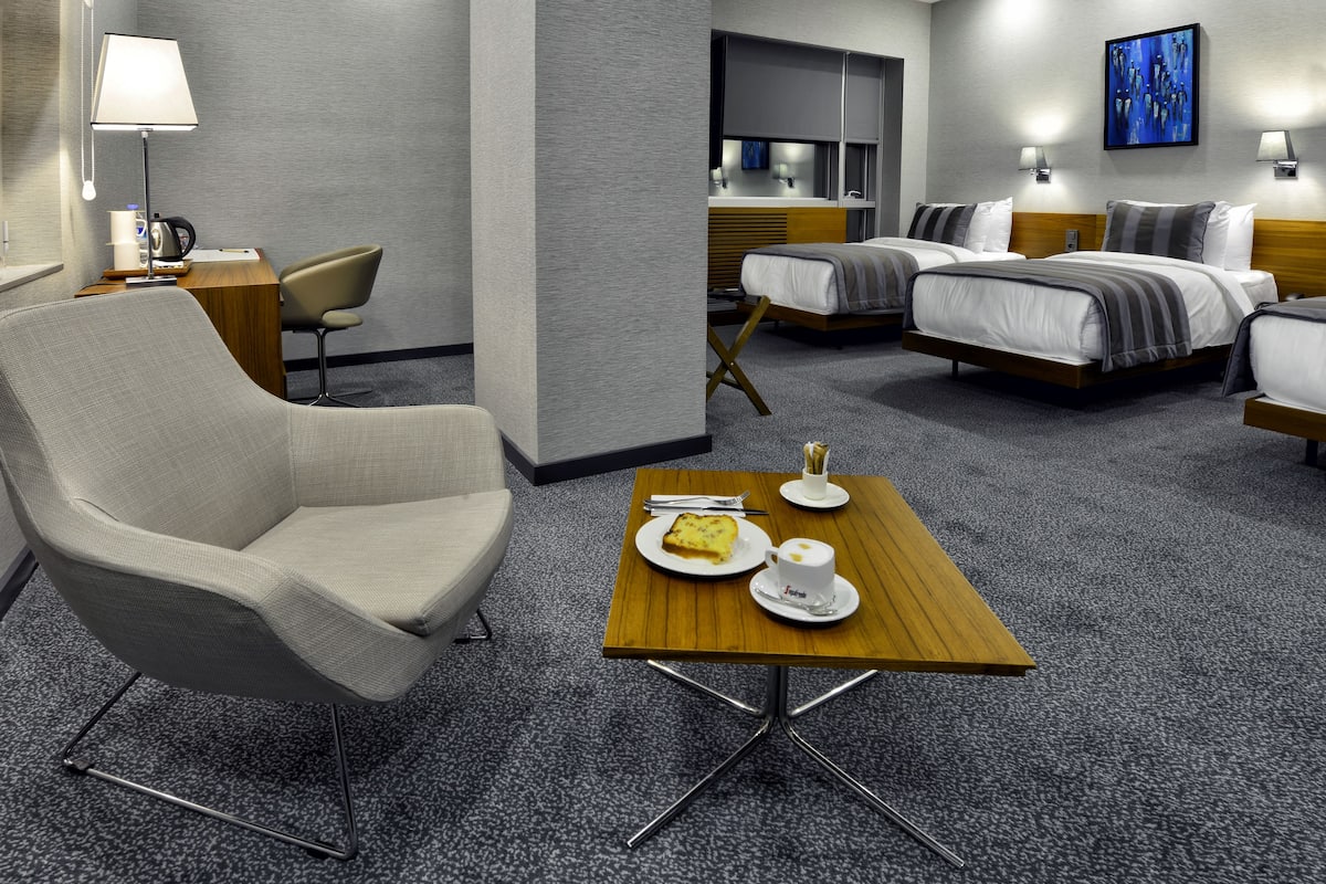 A hotel room with a single bed, a double bed, a seating area with a chair, a small table with a plate of food, and a lamp.