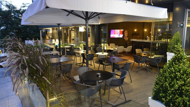Een terras met tafels en stoelen, een grote parasol en een bar met een televisiescherm.