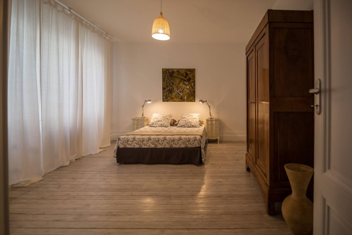 A bedroom with a wooden floor, a bed with floral bedding, two bedside lamps, a hanging light fixture, a wooden wardrobe, and a painting on the wall.