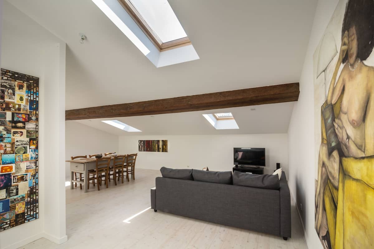 A spacious living area with a gray sofa, wooden dining table, and chairs. There are skylights on the ceiling, a painting on the wall, and a television on a stand.