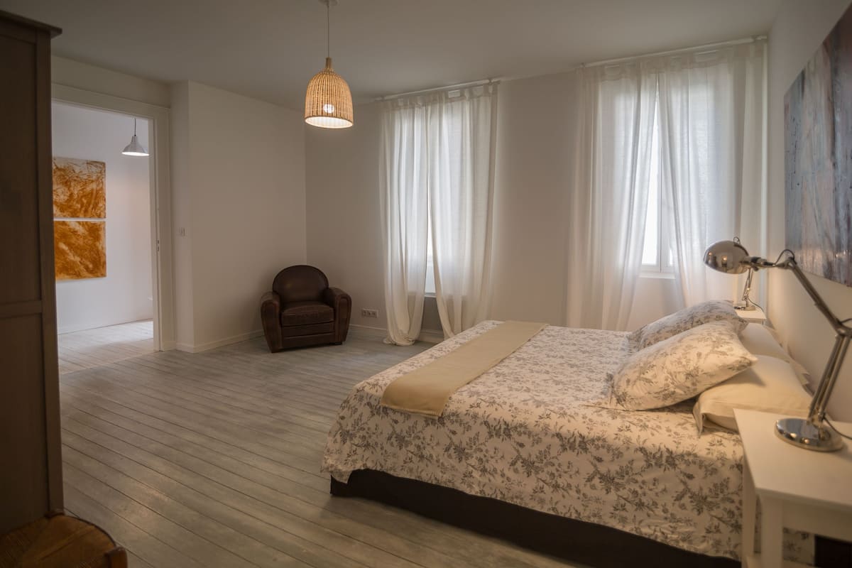 A bedroom with a bed, a chair, a desk with a lamp, and a painting on the wall.