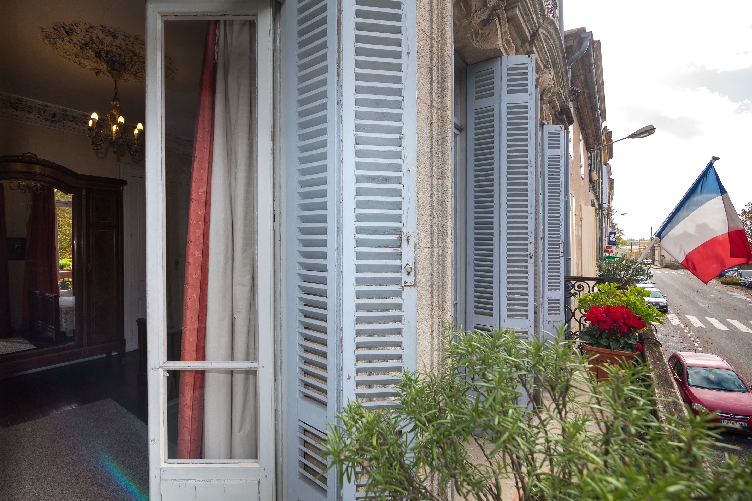 A view from a balcony overlooking a street with a French flag, shutters, and a red car.