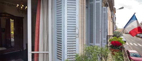 A view from a balcony overlooking a street with a French flag, shutters, and a red car.