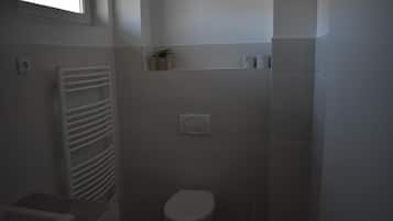Et lille, svagt oplyst badeværelse med toilet, et håndklædetørrestativ og en lille hylde med en plante.
