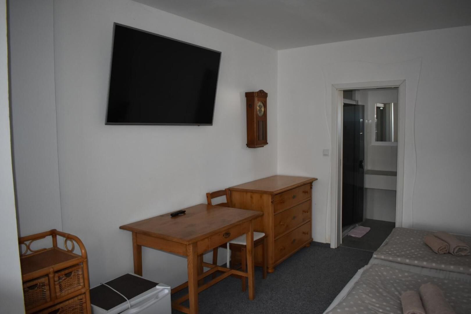 Une pièce comprenant un bureau en bois, un téléviseur fixé au mur, une commode en bois, une chaise en osier et un petit réfrigérateur.