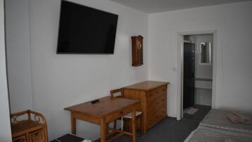 Une pièce comprenant un bureau en bois, un téléviseur fixé au mur, une commode en bois, une chaise en osier et un petit réfrigérateur.