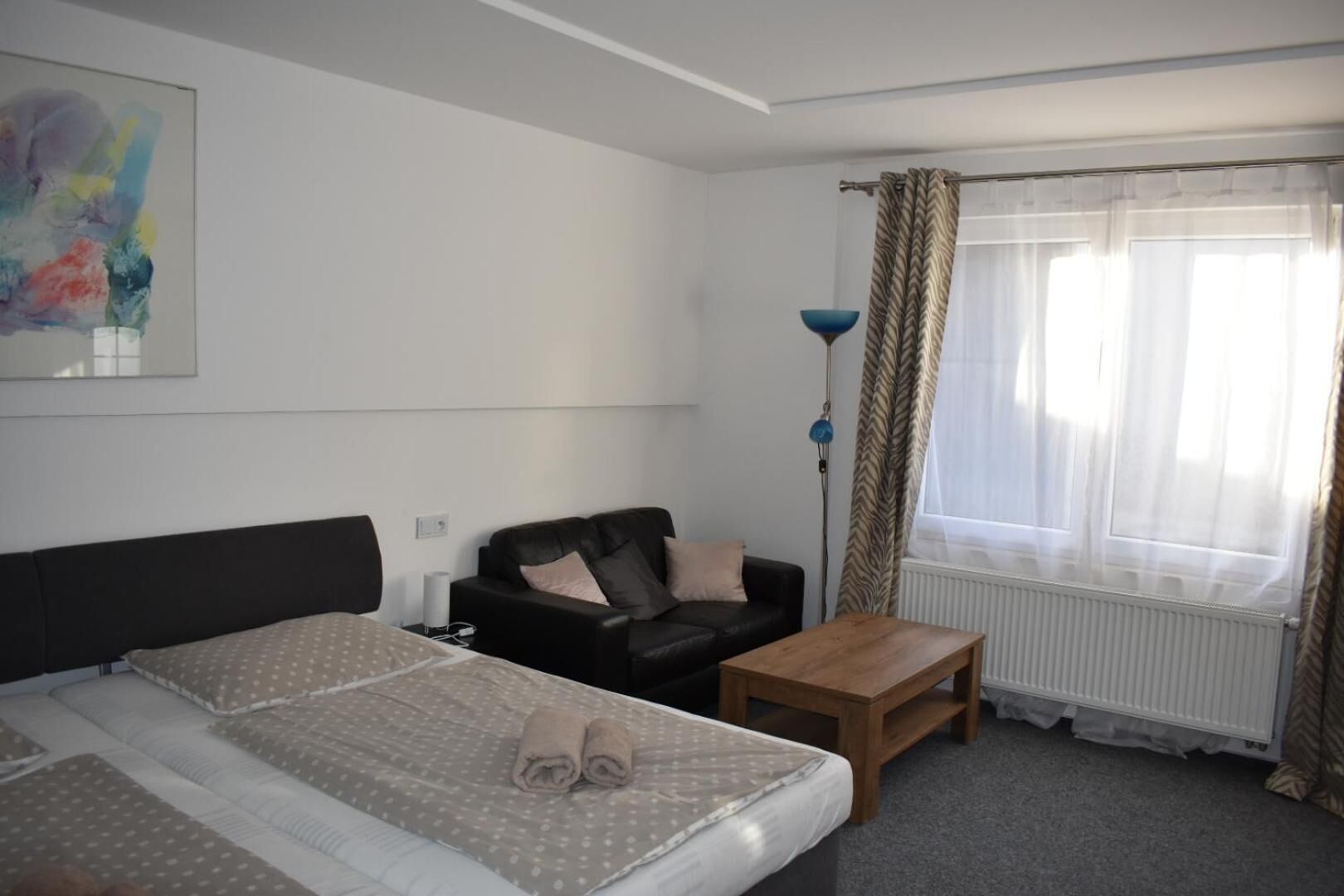 Une chambre d’hôtel avec un lit, un canapé, une table en bois, une lampe sur pied et une fenêtre avec des rideaux légers.