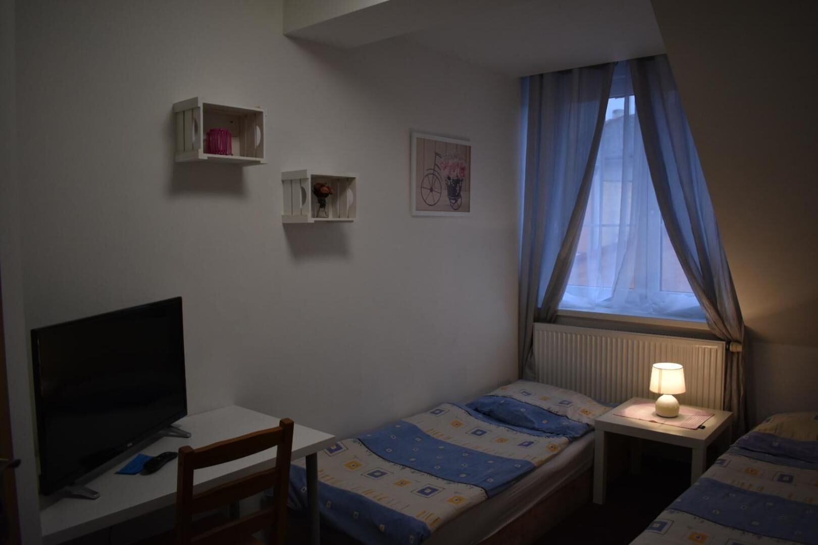 Een kleine, eenvoudige kamer met een bed, een bureau en een televisie. Er zijn twee nachtkastjes met lampen, en een raam met gordijnen.