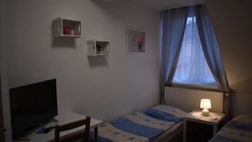 Een kleine, eenvoudige kamer met een bed, een bureau en een televisie. Er zijn twee nachtkastjes met lampen, en een raam met gordijnen.
