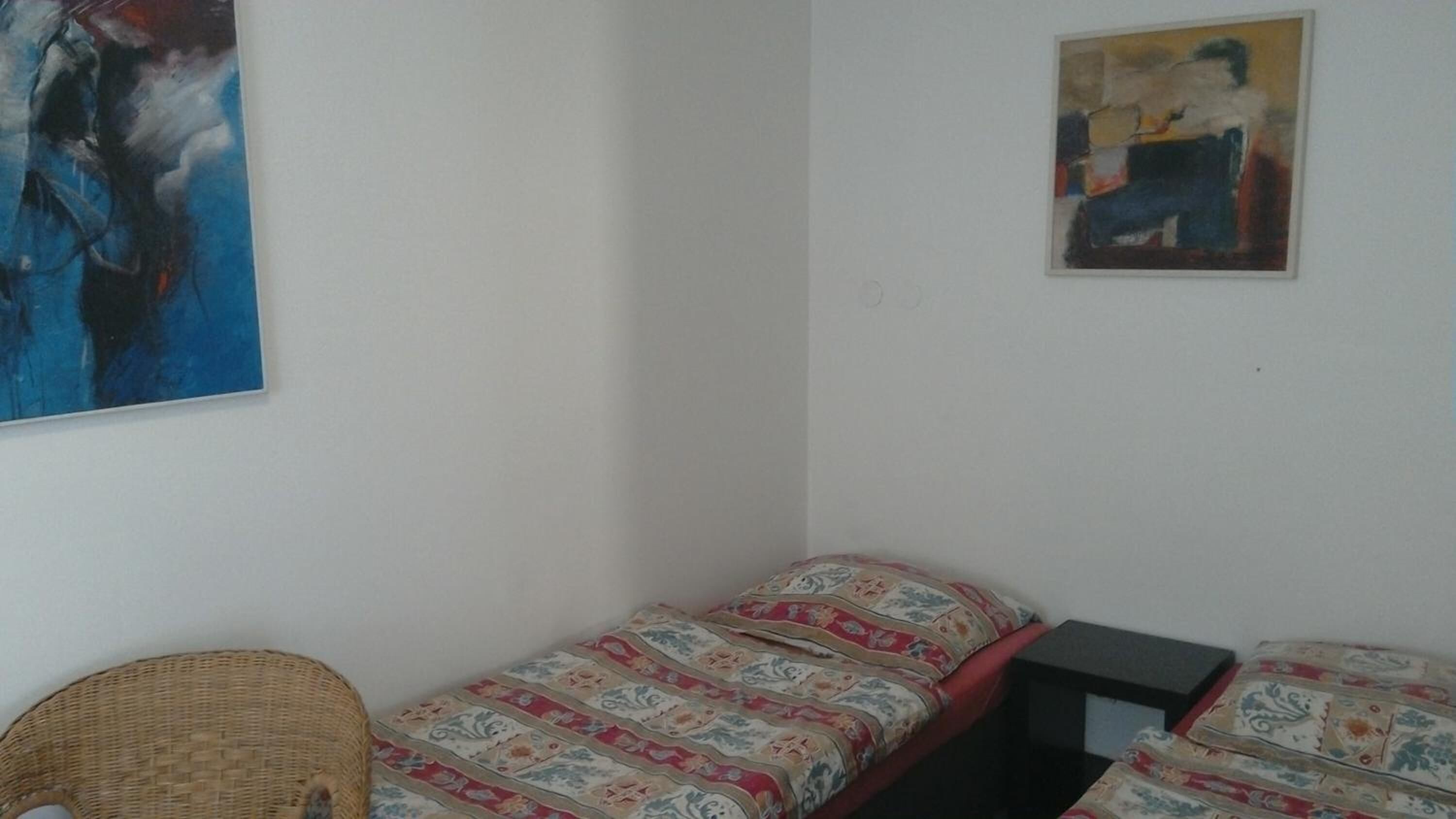 A small, simple room with two single beds, a wicker chair, and two abstract paintings on the walls.