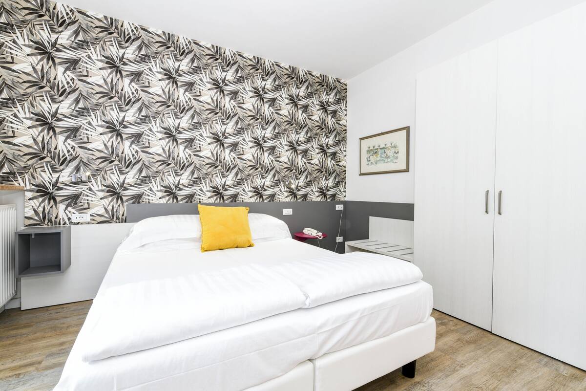 Ein modernes Schlafzimmer mit Wandmuster, einem Bett mit weißer Bettwäsche und einem gelben Kissen, einem Nachttisch, einem Kleiderschrank und einem gerahmten Bild an der Wand.