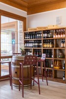 Una zona de bar con estanterías de madera llenas de botellas de vino, dos taburetes y una mesa de cristal.