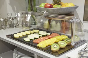 A buffet with a variety of fruits and meats, including sliced apples, oranges, kiwi, and salmon, displayed under a protective glass cover. - Welcome Gros (San Sebastián)