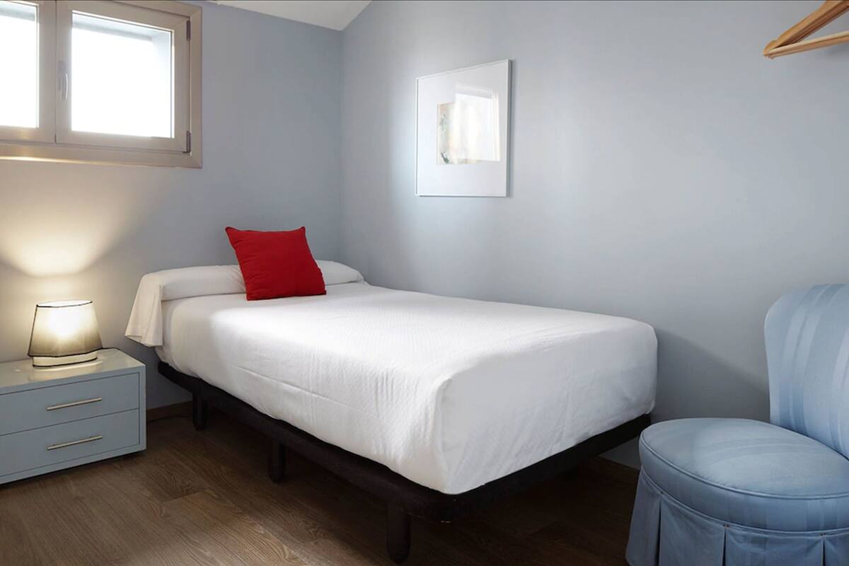 Una cama bien hecha con edredón blanco y almohada roja, una lámpara de noche, un cajón pequeño, un cuadro en la pared y una silla azul.