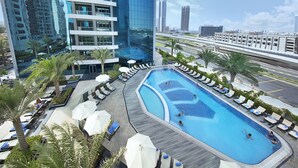 Ein Pool auf dem Dach mit Liegestühlen und Sonnenschirmen, umgeben von Palmen und modernen Gebäuden.