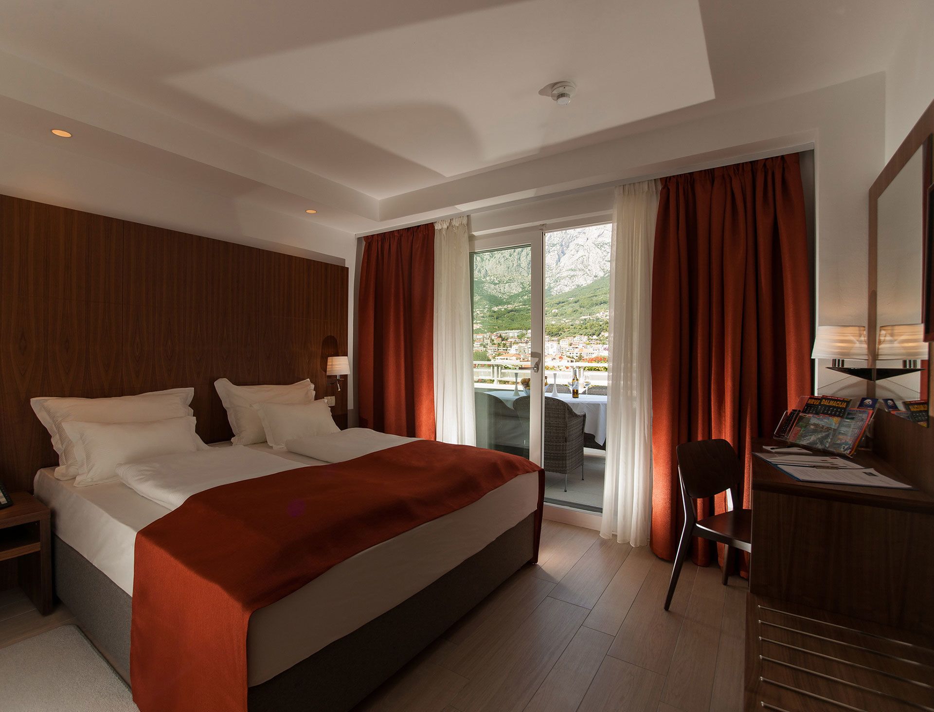 A hotel room with a large bed, a desk with a chair, and a view of a mountainous landscape through the window.