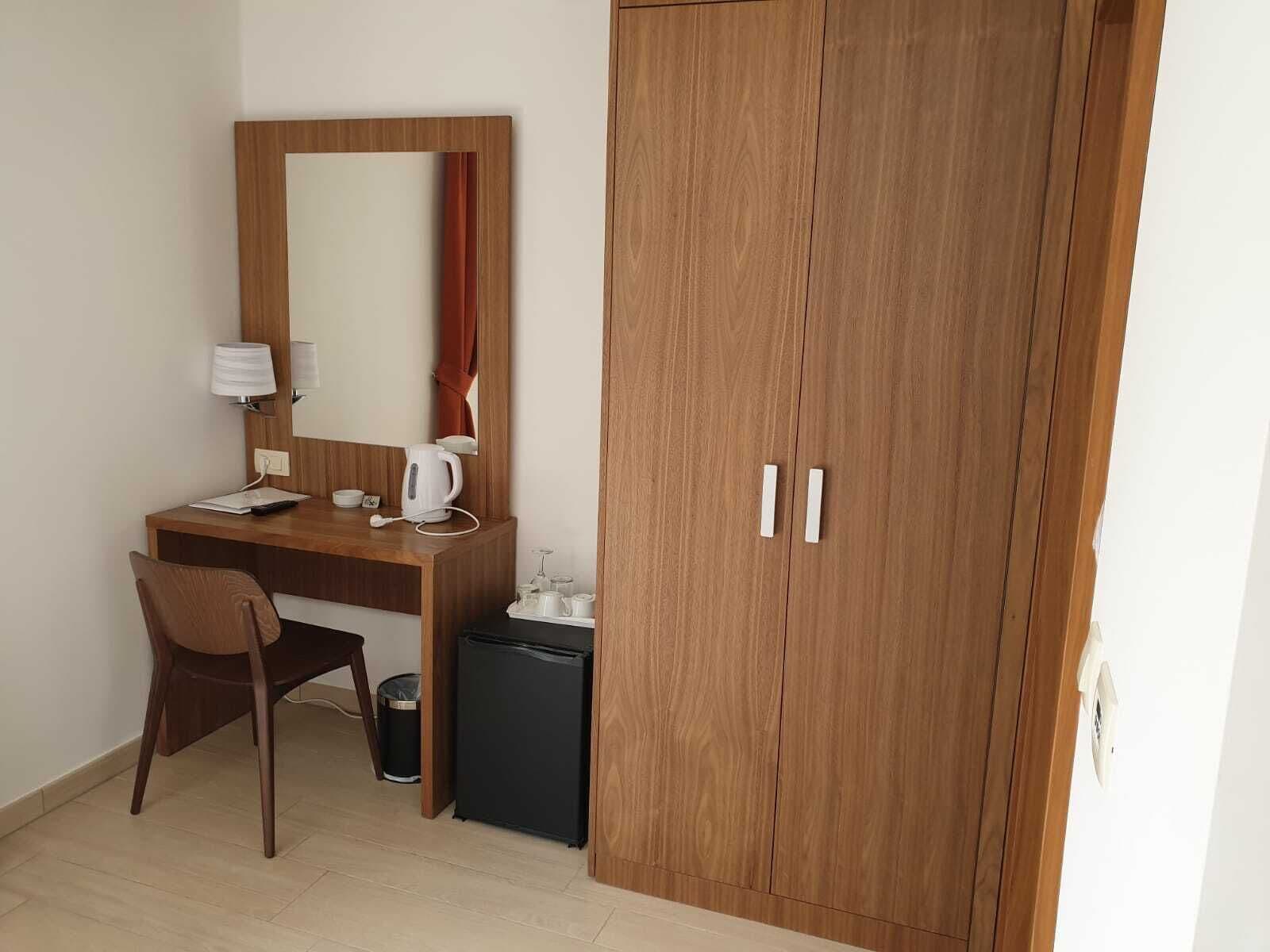 Een hotelkamer met een houten bureau, een stoel, een spiegel, een kledingkast en een kleine koelkast.