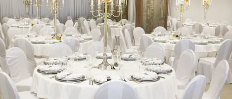 Een balzaal met ronde tafels gedekt voor een formeel evenement, elk met witte tafellakens, bestek en bloemstukken als decoratie.
