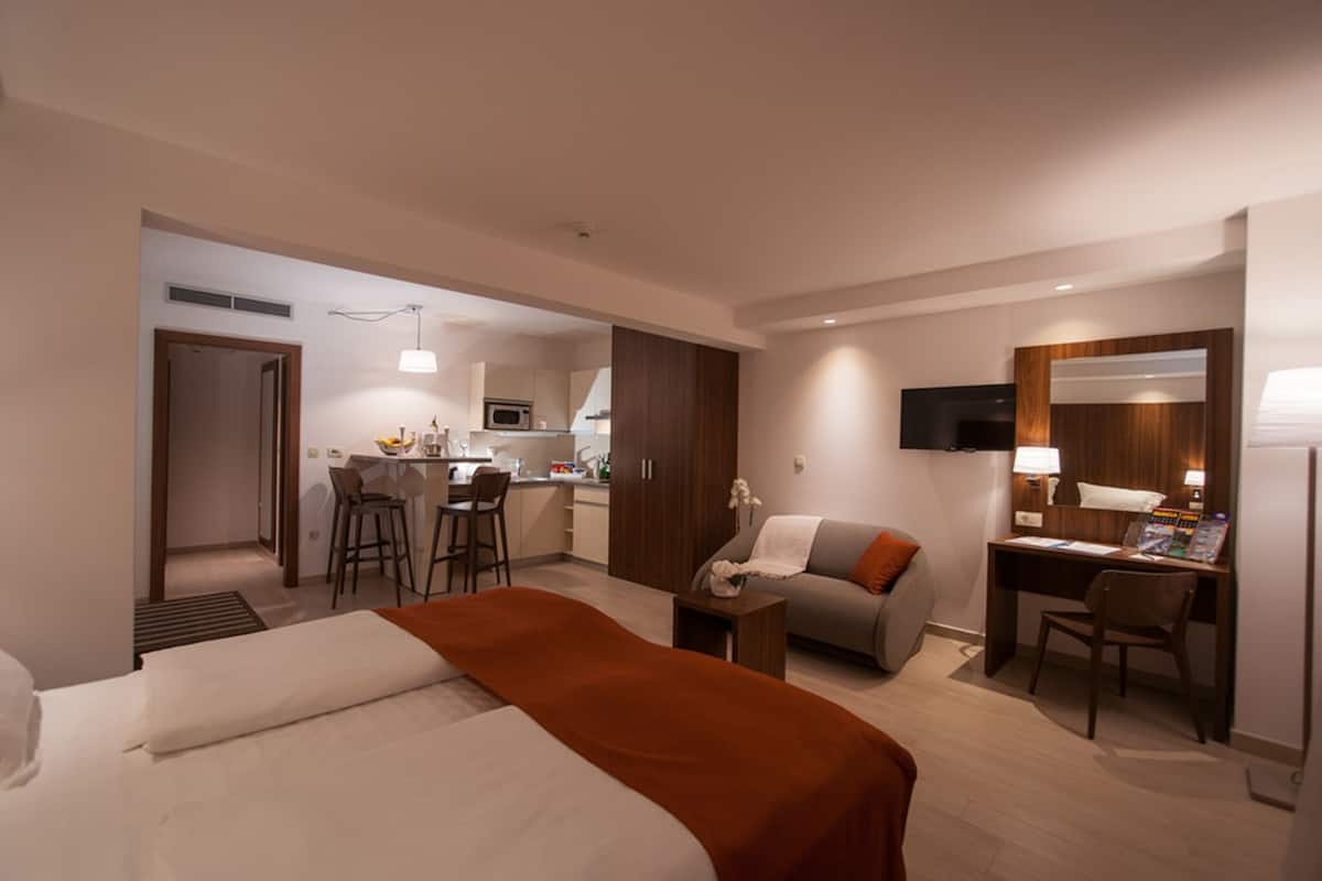 Moderni hotellihuone, jossa on sänky, työpöytä, tuoli, televisio ja taustalla pieni keittiönurkkaus.