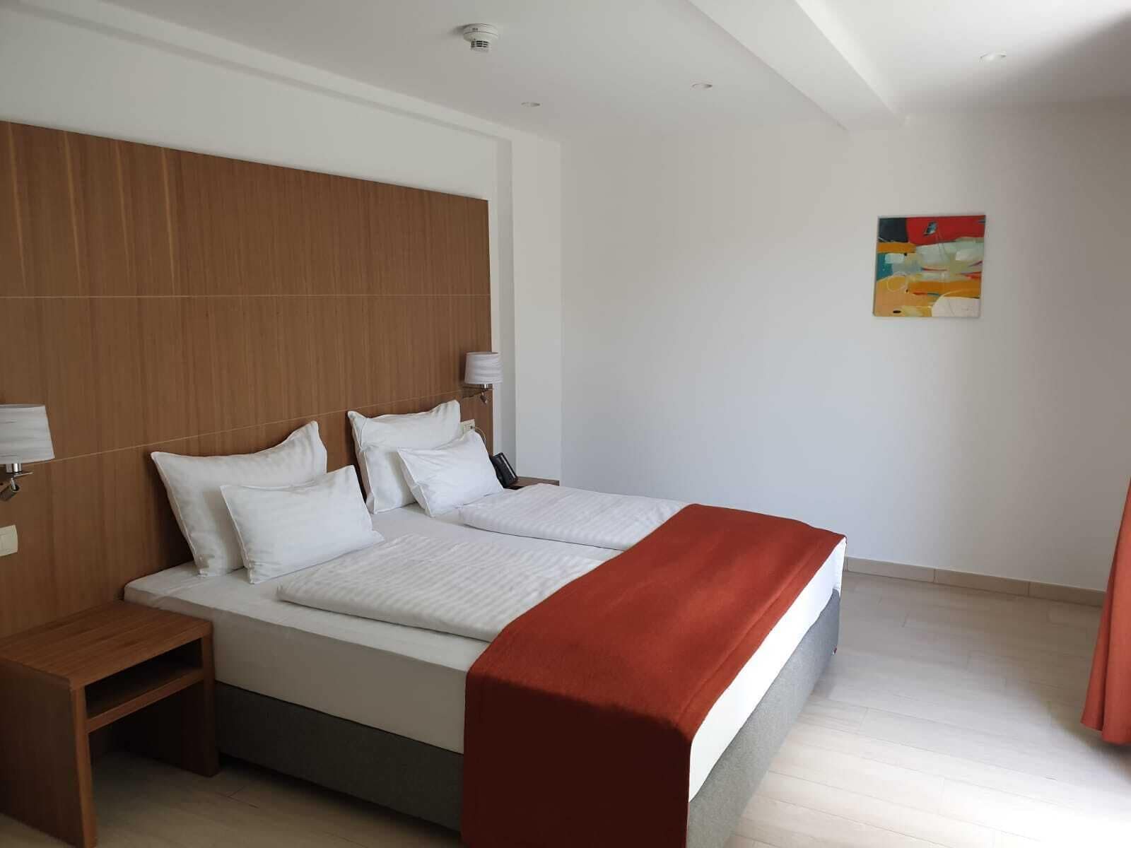 A hotel room with a large bed, wooden headboard, two bedside tables, a red throw blanket, and a single wall-mounted lamp.
