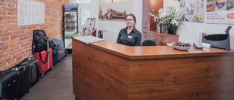 A reception desk with a wooden counter, a brick wall, and a chandelier. There are suitcases and a refrigerator in the background.