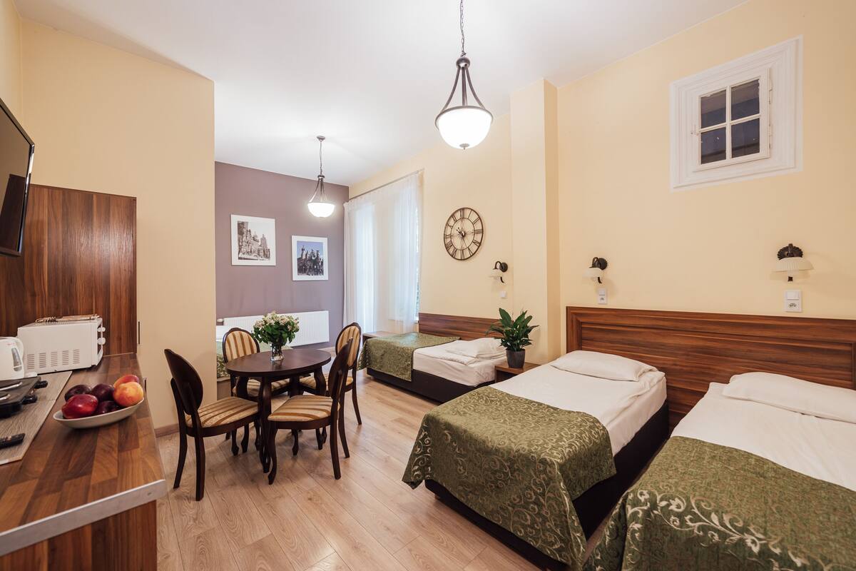 Habitación de hotel con dos camas, una mesa de comedor con sillas, una pequeña cocina y un ventanal con cortinas.