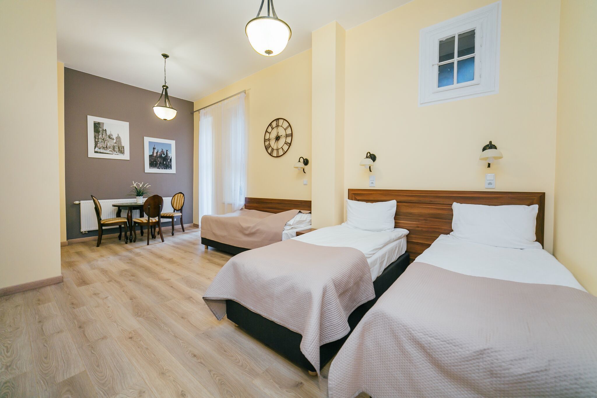 A hotel room with two beds, a dining table, chairs, a clock, and a window.