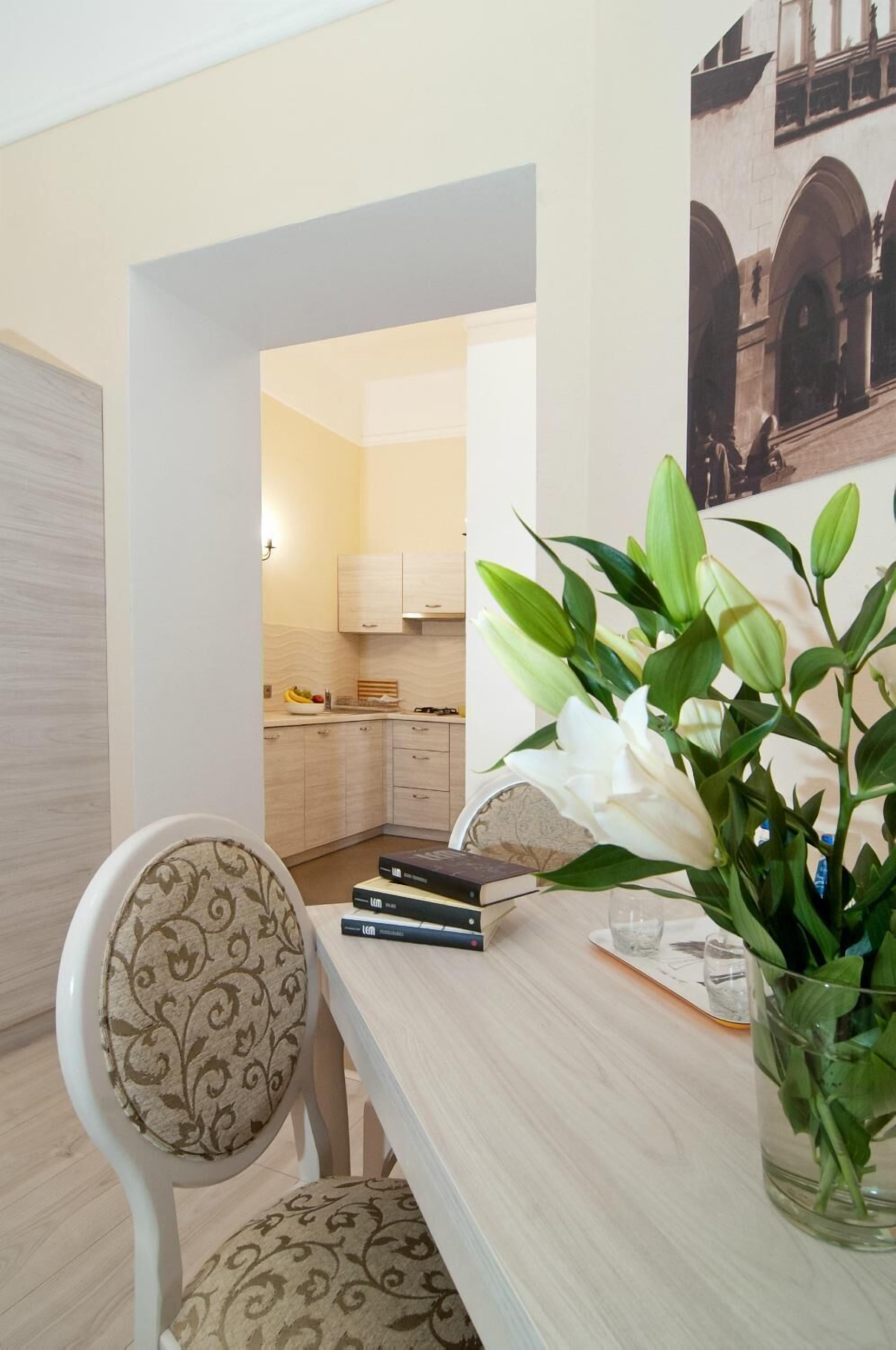 A dining area with a table, chairs, and a vase of flowers. There is a kitchen visible in the background.
