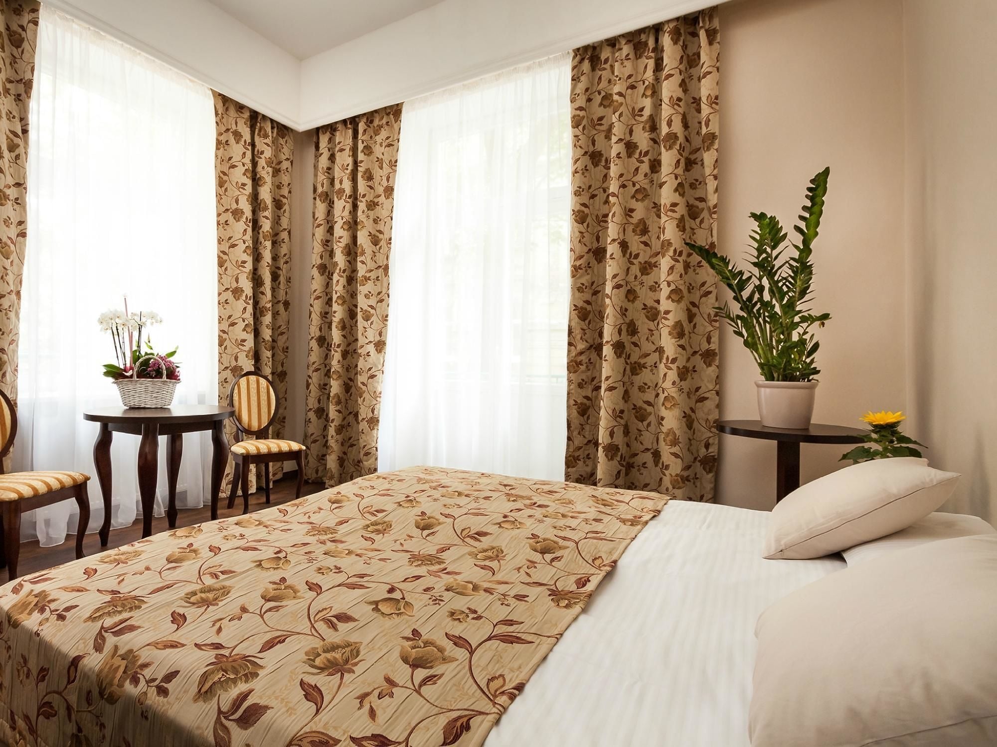 A hotel room with a large bed, a small round table with a chair, a potted plant, and a window with curtains.