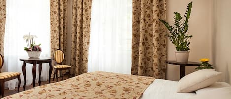 Quarto de hotel com uma cama grande, uma mesinha redonda com uma cadeira, uma planta em vaso e uma janela com cortinas.