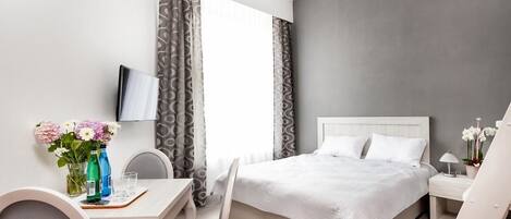 A modern hotel room with a bed, a desk with chairs, a TV, and a window with curtains.