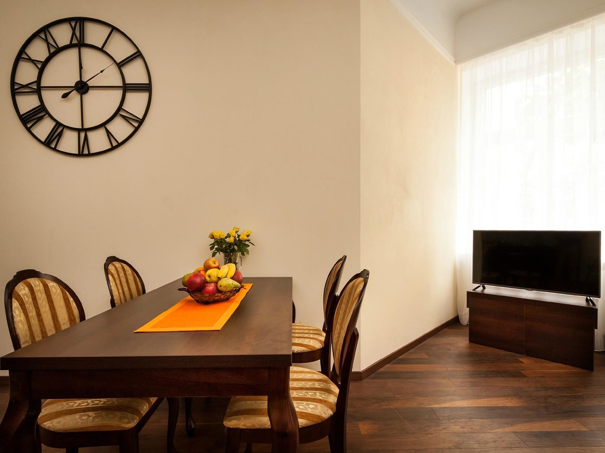 Uma sala de jantar com uma mesa de madeira, quatro cadeiras, uma cesta de frutas e um relógio de parede.