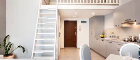 A modern bedroom with a loft-style sleeping area, a staircase, a dining area with a table and chairs, and a kitchen with cabinets and appliances.