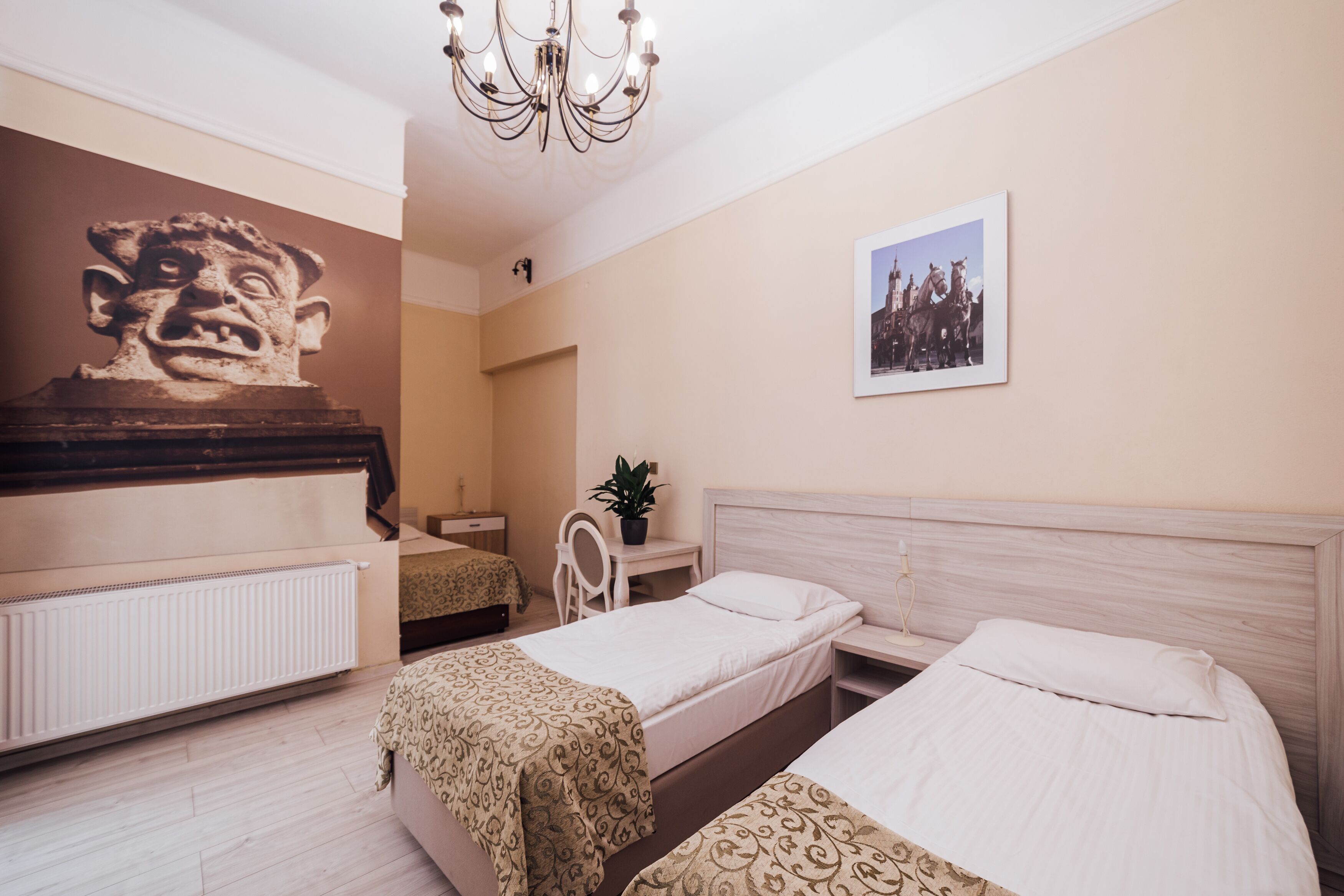A hotel room with two single beds, a wooden headboard, a radiator, a desk with a chair, a potted plant, a wall-mounted picture, and a large sculpture.