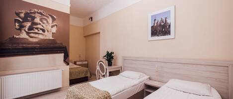 A hotel room with two single beds, a wooden headboard, a radiator, a desk with a chair, a potted plant, a wall-mounted picture, and a large sculpture.