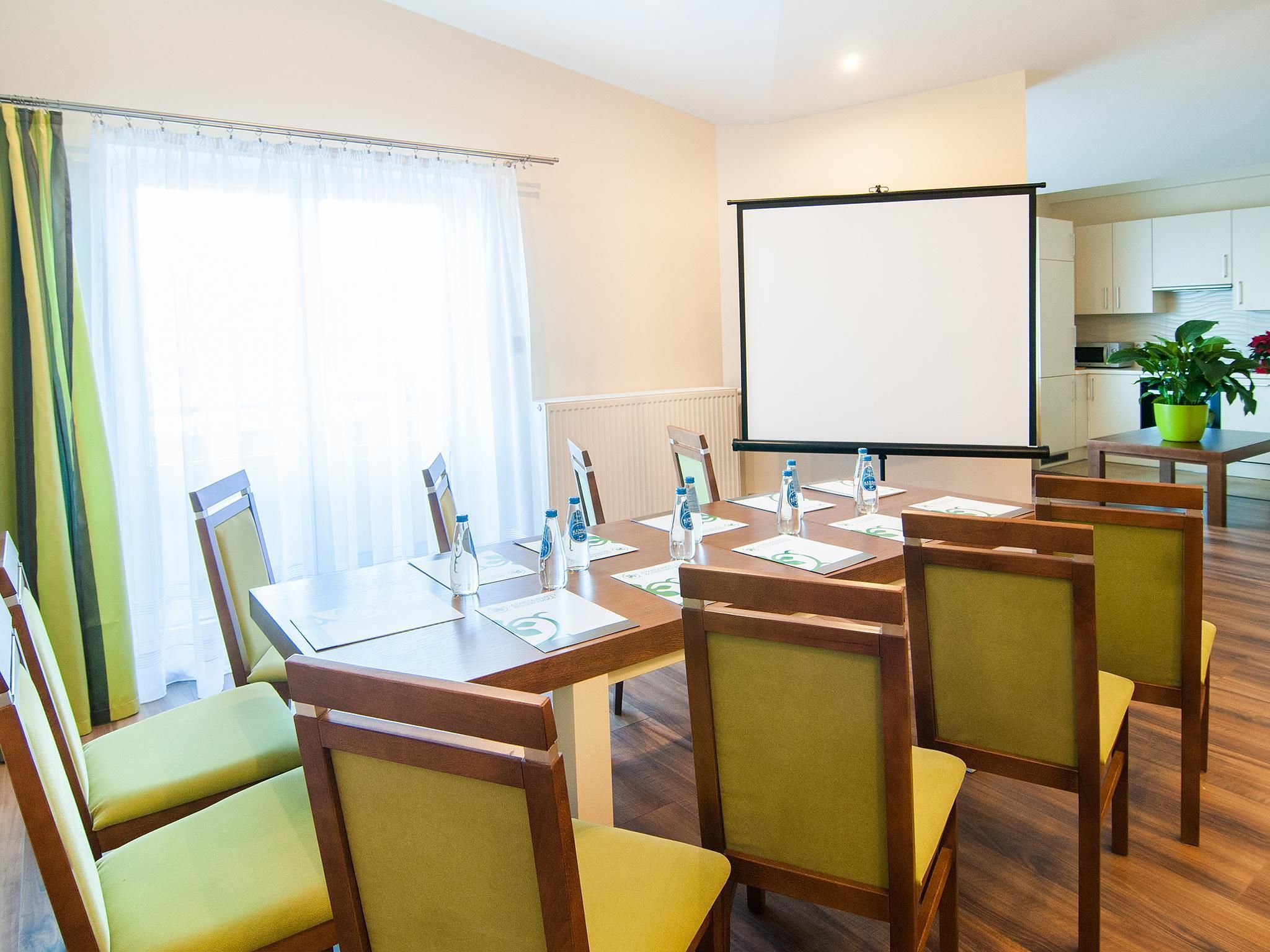 Una sala de conferencias con una mesa grande, sillas, una pantalla de proyector y un ventanal con cortinas.