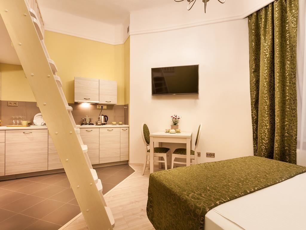A compact hotel room with a bed, a desk, a chair, a television mounted on the wall, and a kitchen area with cabinets and a stove.