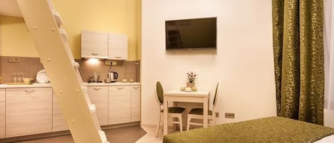 Habitación de hotel compacta con cama, escritorio, silla, televisor montado en la pared y zona de cocina con armarios y estufa.