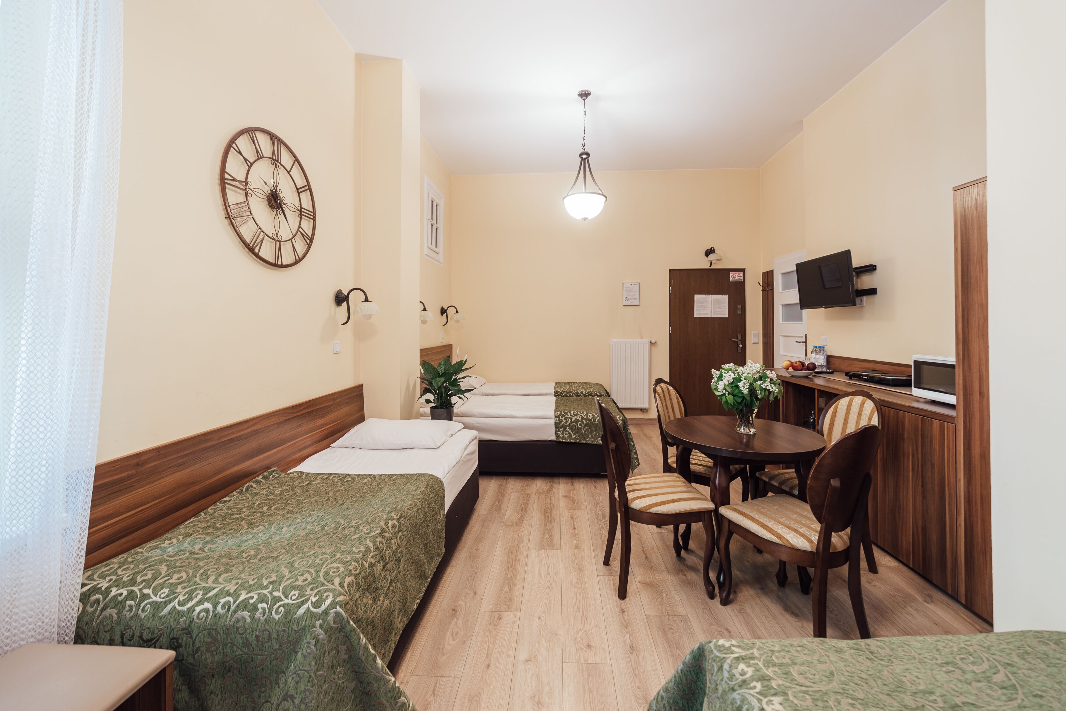 Ein gemütliches Hotelzimmer mit zwei Einzelbetten, einem kleinen Esstisch mit Stühlen, einer Wanduhr, einem Fernseher und einer Heizung.