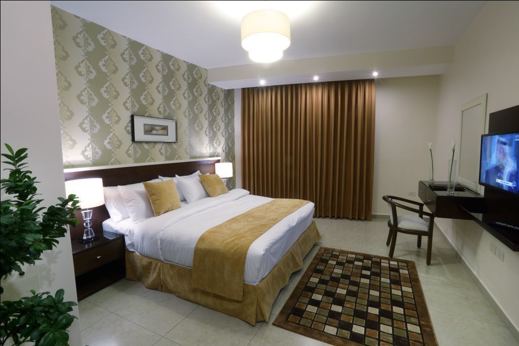 Quarto de hotel com cama, mesa de cabeceira, luminária, televisão e escrivaninha com cadeira.