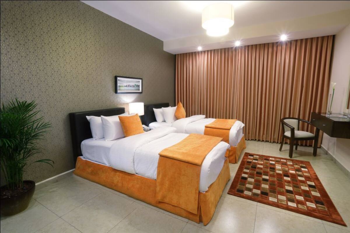 A hotel room with two beds, a desk, a chair, a potted plant, and a patterned rug.