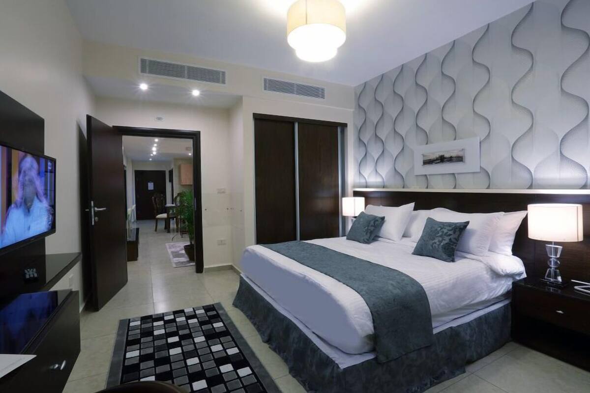A modern hotel room with a large bed, a TV, a desk, and a patterned wall.