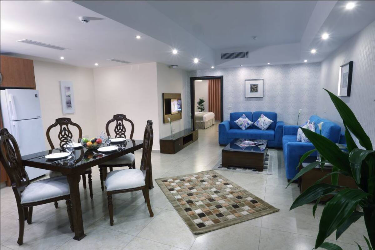 A modern living room with a blue sofa, a dining table with chairs, and a kitchen area with a refrigerator.