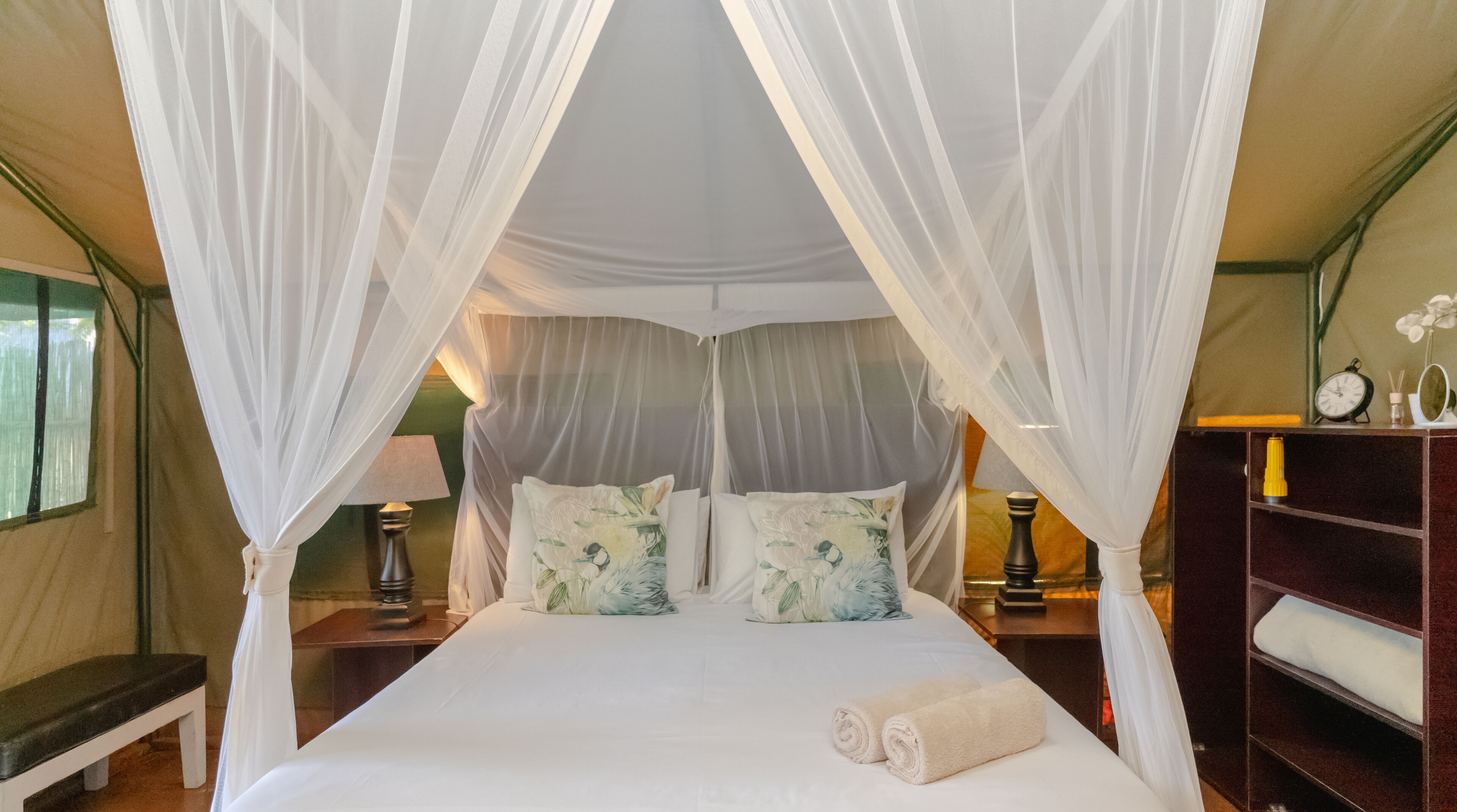 A four-poster canopy bed with white linens, two nightstands with lamps, and a bedside table with a clock and a bottle.