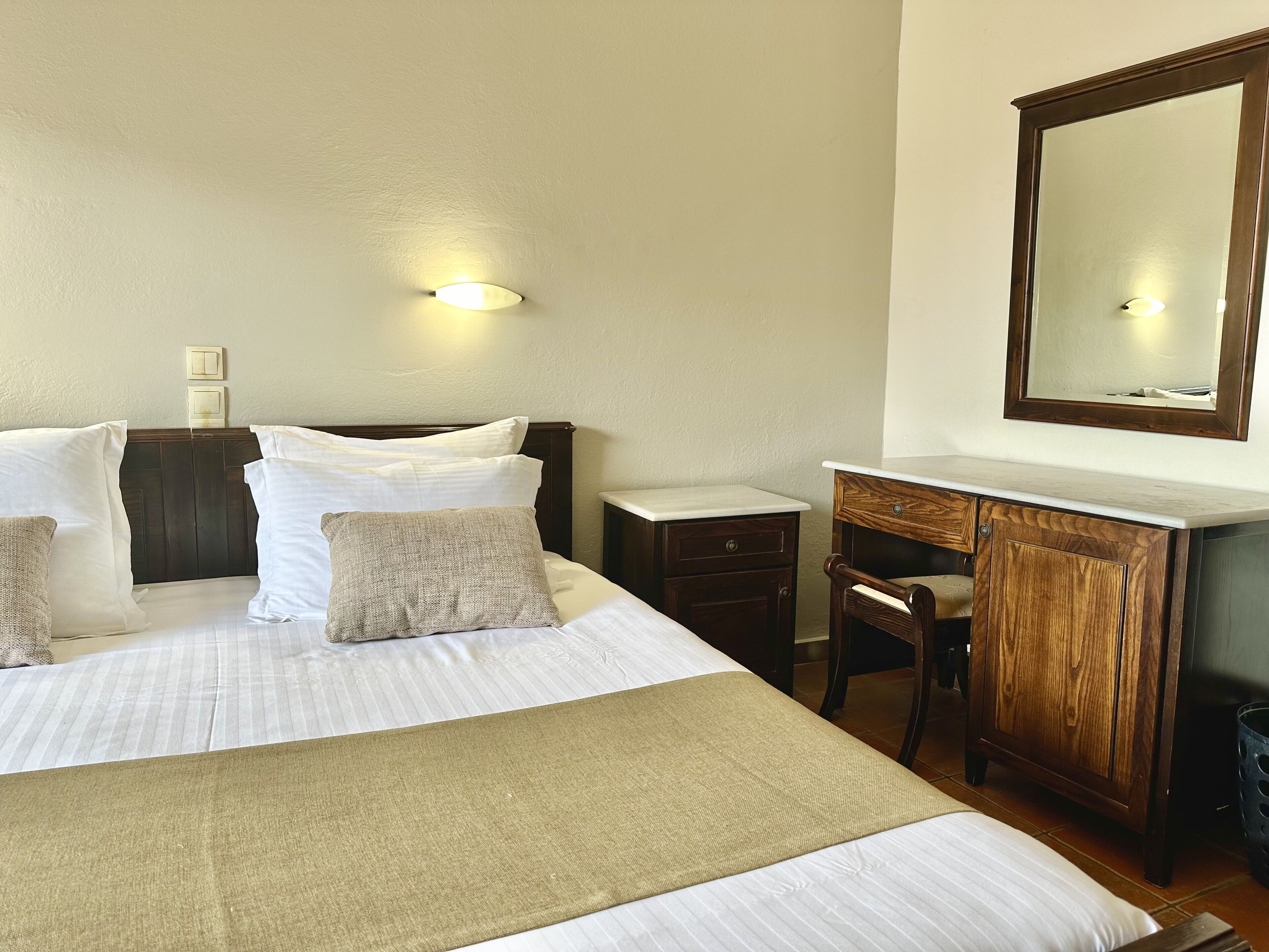 Habitación de hotel con cama, mesitas de noche, cómoda de madera, espejo y lámpara de pared.