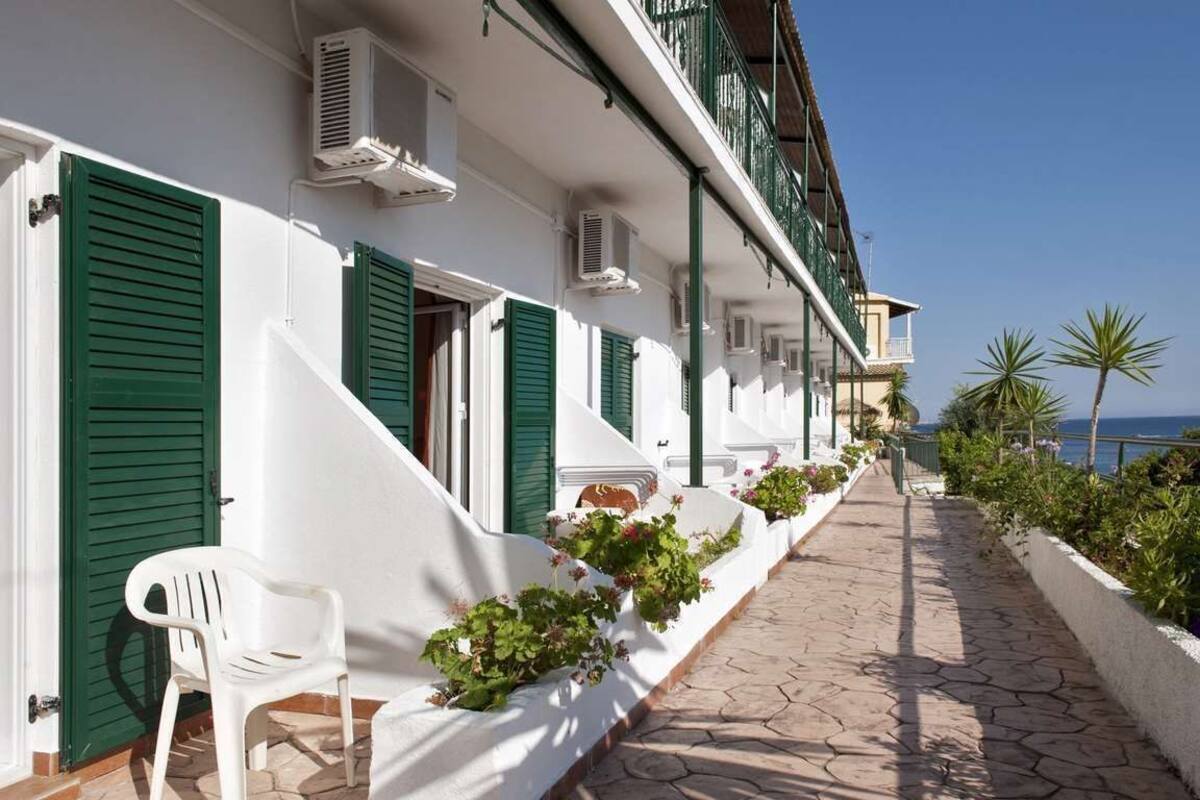 A white building with green shutters, air conditioning units, and a pathway lined with potted plants.