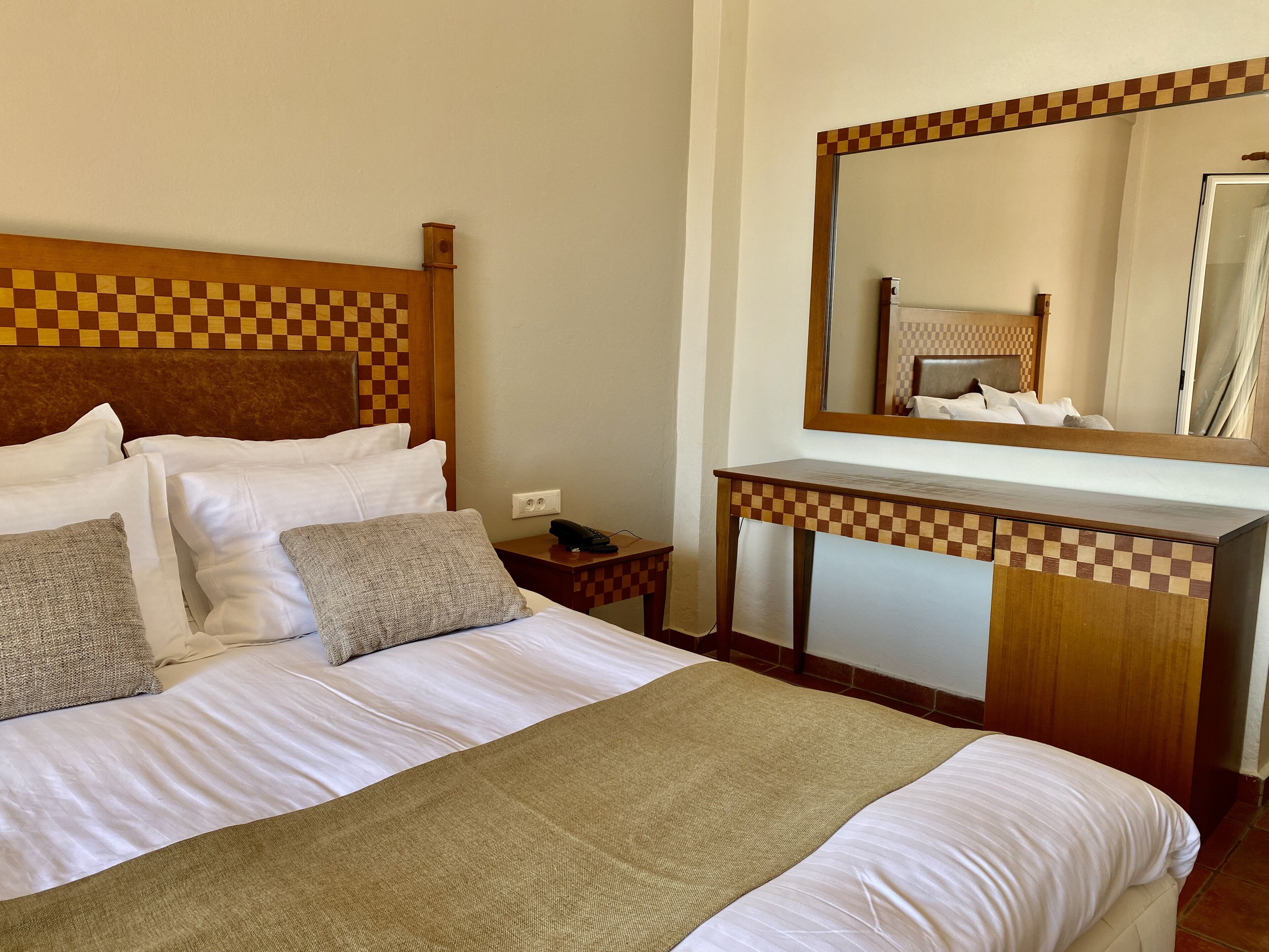 Ein Hotelzimmer mit kariertem Kopfteil, einem großen Spiegel über einem Holzkonsolentisch und einem Bett mit weißen Bettwäsche und einer beigefarbenen Tagesdecke.