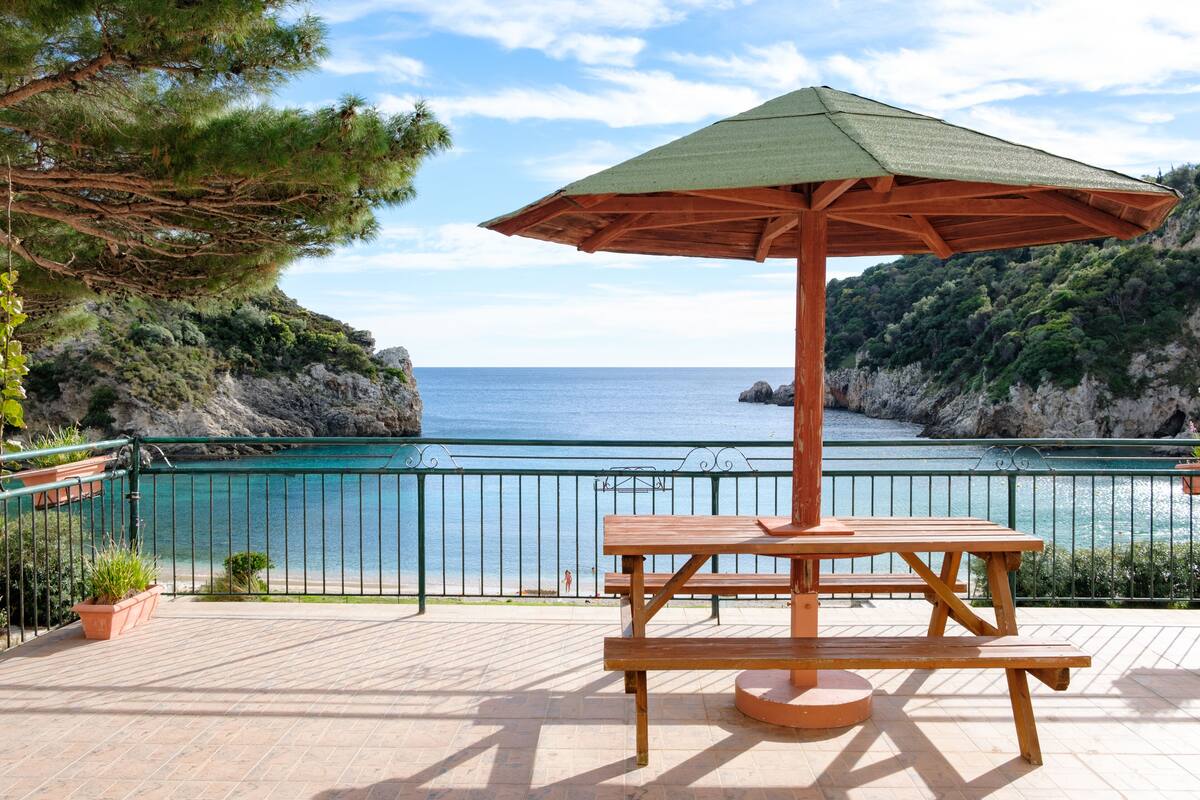 A wooden picnic table with a green umbrella on a tiled patio overlooking a coastal area with a clear view of the sea and rocky cliffs.