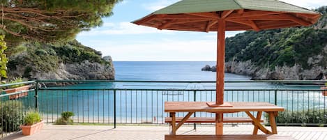 A wooden picnic table with a green umbrella on a tiled patio overlooking a coastal area with a clear view of the sea and rocky cliffs.
