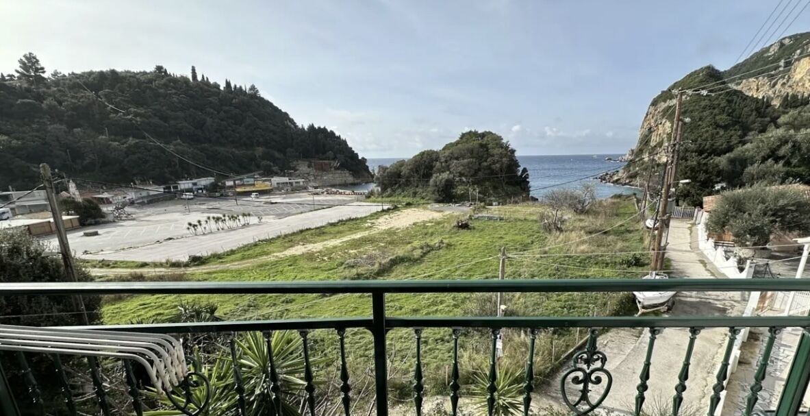 Vista costera desde un balcón con baranda de metal, sobre un estacionamiento y un área verde con árboles y colinas al fondo.